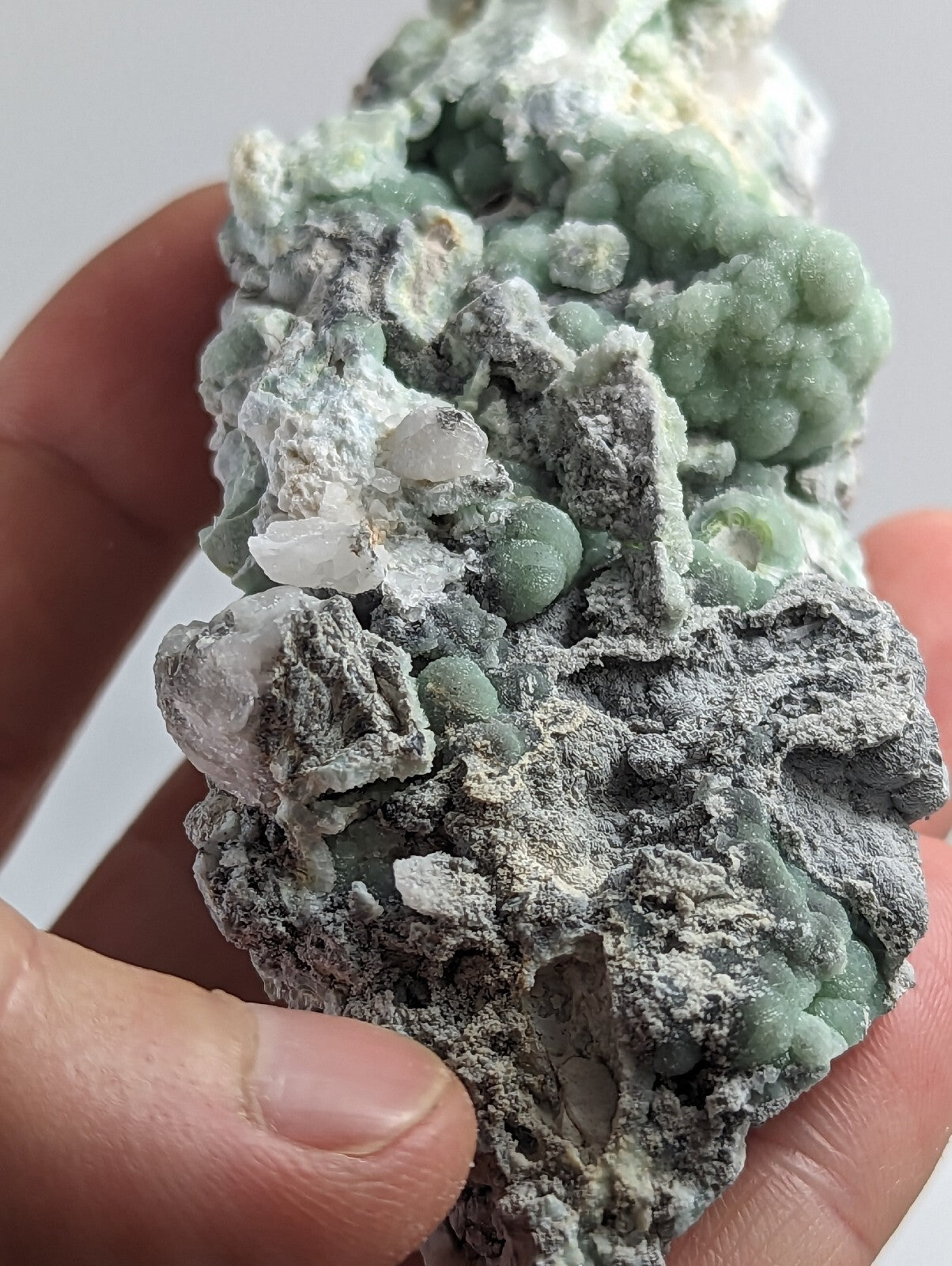 A hand holds a natural mineral specimen featuring clusters of pale green, bead-like crystals embedded in a grayish matrix with white crystalline patches.