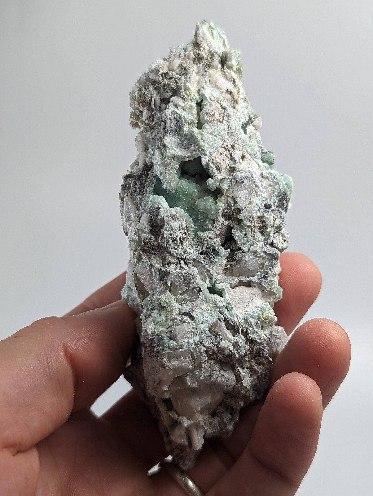 A hand holds a rough, irregularly shaped mineral specimen featuring patches of translucent light green crystals embedded in a matrix of white and gray material.