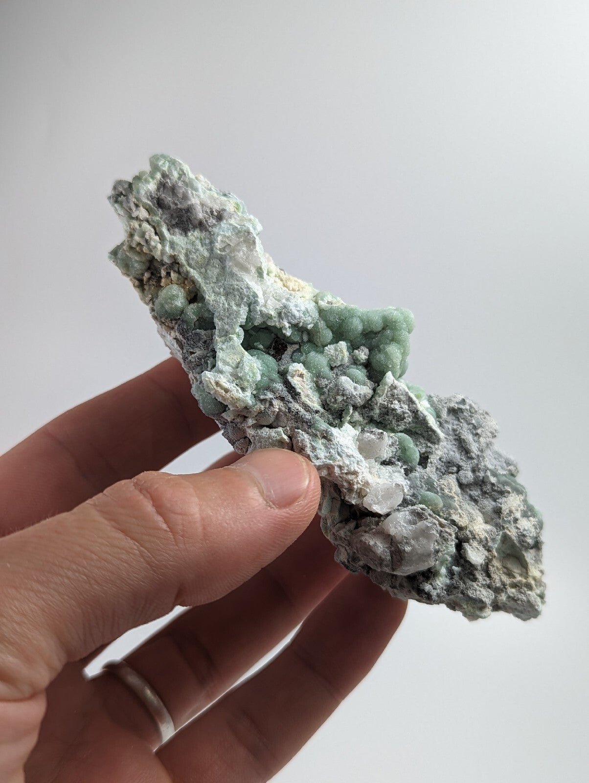 A hand holds a rough, irregularly shaped mineral specimen featuring clusters of pale green crystals embedded in a grayish matrix with white veining.