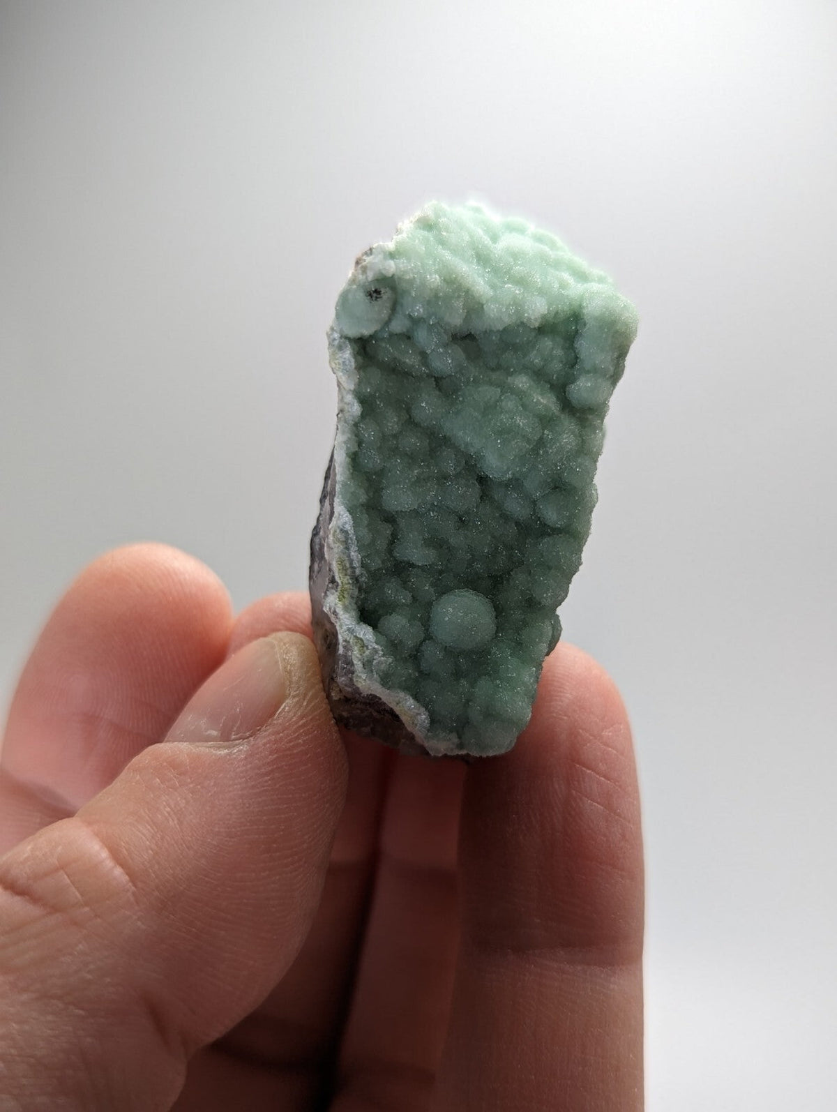 A hand holds a light green, textured mineral specimen with rounded crystalline formations.
