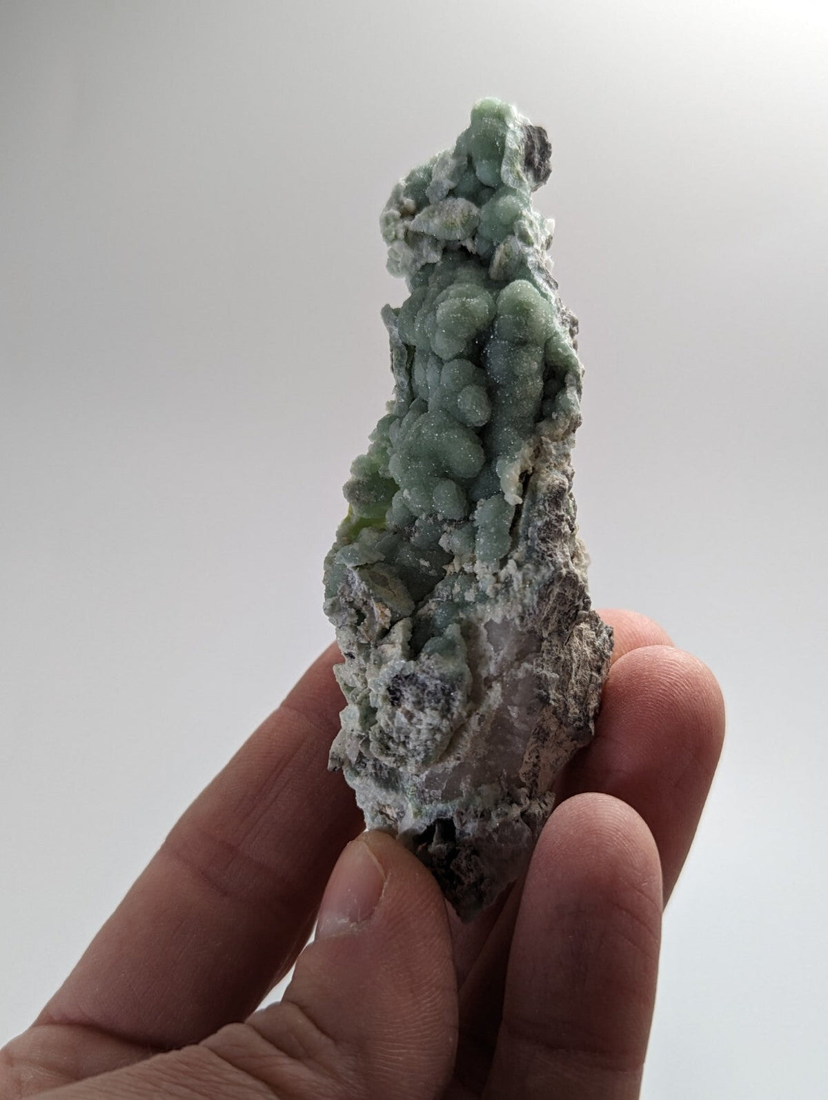 A hand holds a striking green mineral specimen featuring clustered, bead-like formations atop a rougher, grayish base.