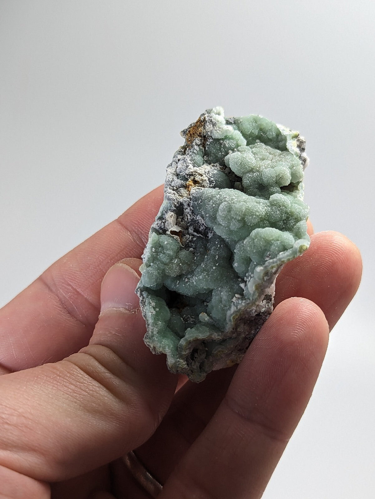 A hand holds a light green, textured mineral specimen with crystalline formations and patches of grayish matrix.