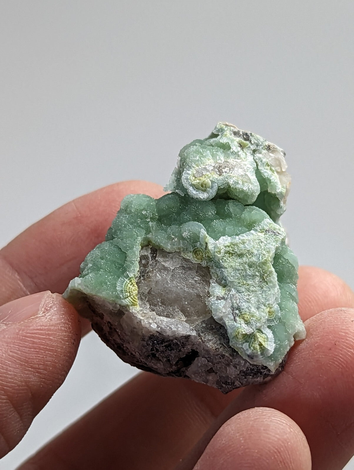A hand holds a rough, green mineral specimen with intricate crystalline formations and patches of white calcite.
