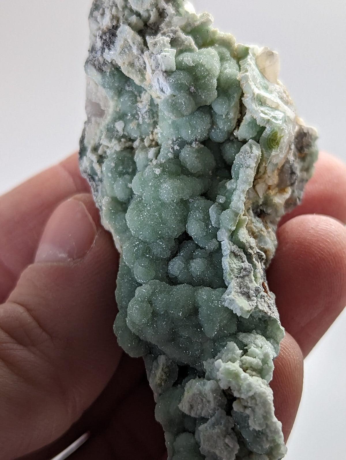 A hand holds a light green, crystalline mineral specimen with rounded, bubbly formations and patches of white and gray matrix.