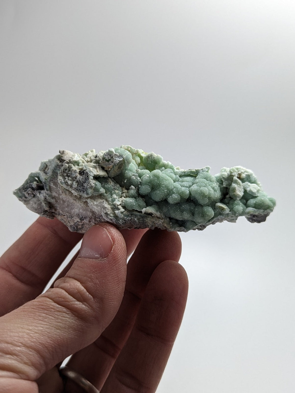 A hand holds a natural mineral specimen featuring clusters of light green, bumpy crystals growing from a darker gray matrix.