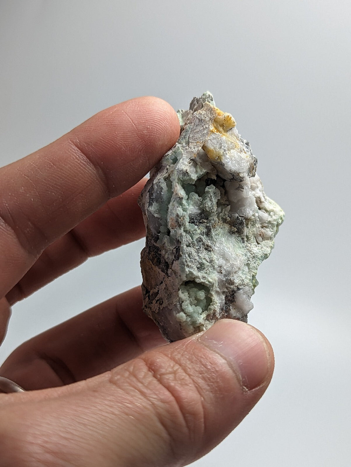 A rough, irregularly shaped mineral specimen with a mottled surface of pale green and white patches accented by small yellow inclusions.