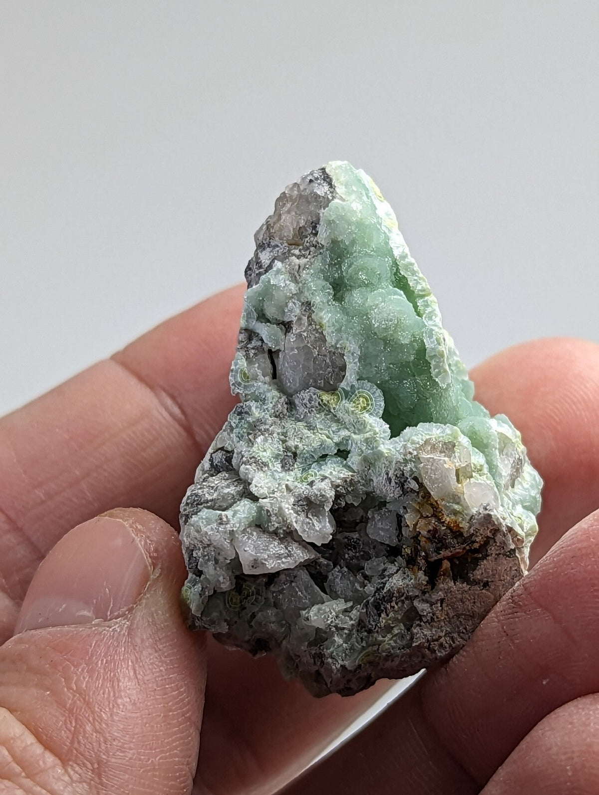 A hand holds a natural green mineral specimen with crystalline formations and earthy gray matrix.