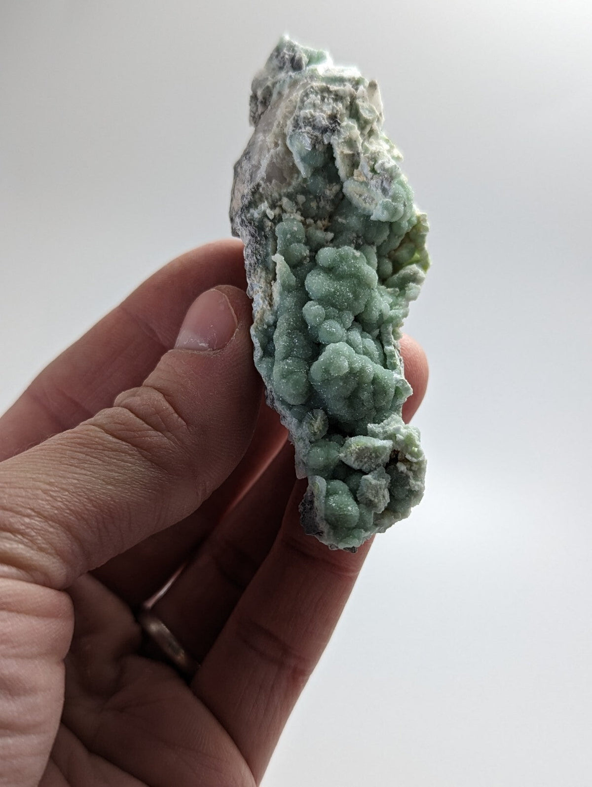 A hand holds a natural mineral specimen featuring vibrant green, clustered crystals growing from a rougher gray matrix.