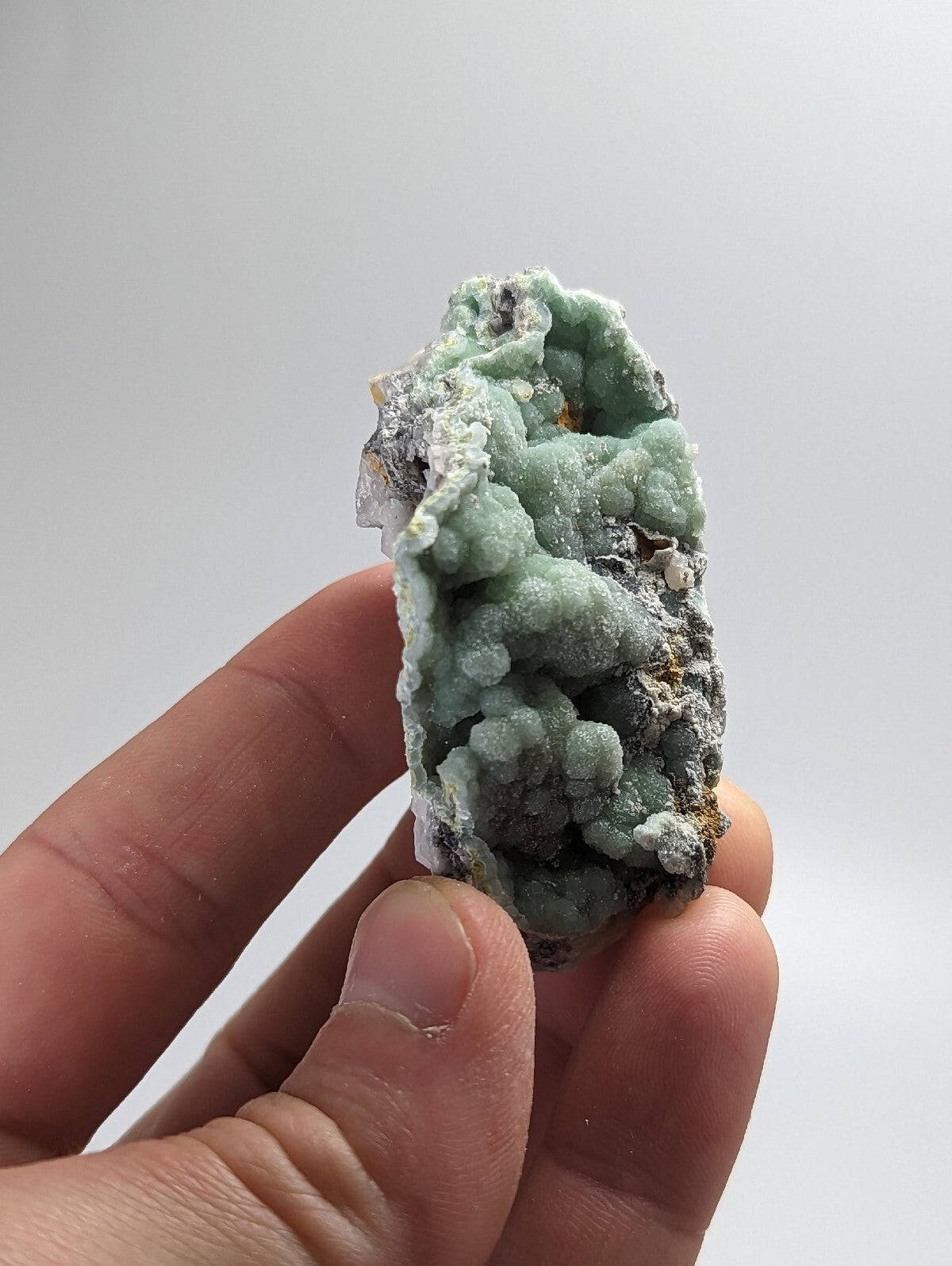 A hand holds a light green, textured mineral specimen with crystalline formations and patches of gray matrix.