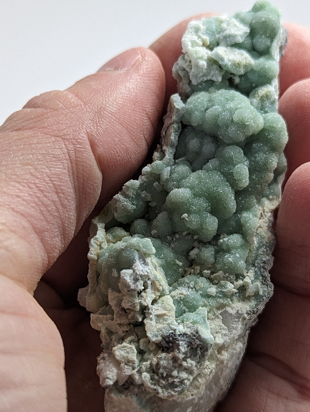 A hand holds a light green, bumpy mineral specimen with white and dark gray matrix.