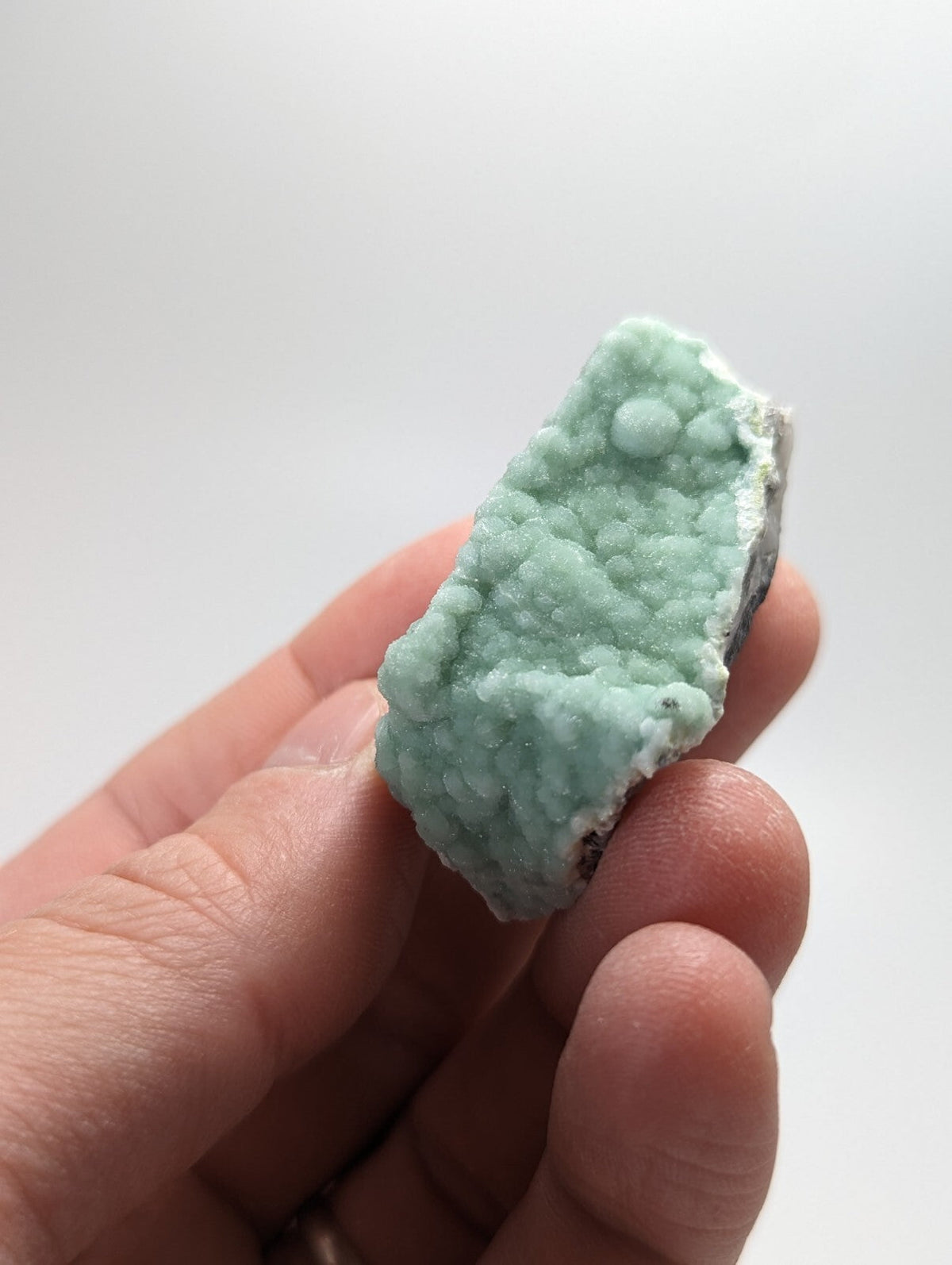 A hand holds a small, irregularly shaped piece of light green mineral with a textured surface featuring rounded nodules and some white crystalline areas.