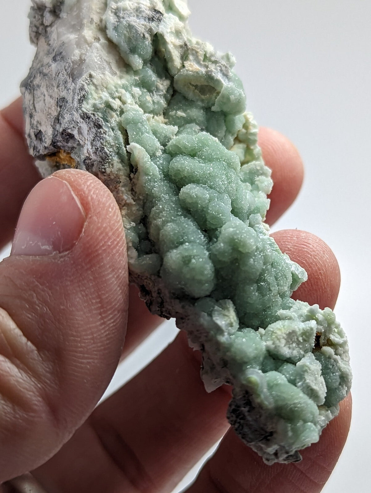 A hand holds a specimen of light green, crystalline mineral formations with rough, textured surfaces and patches of grayish matrix.