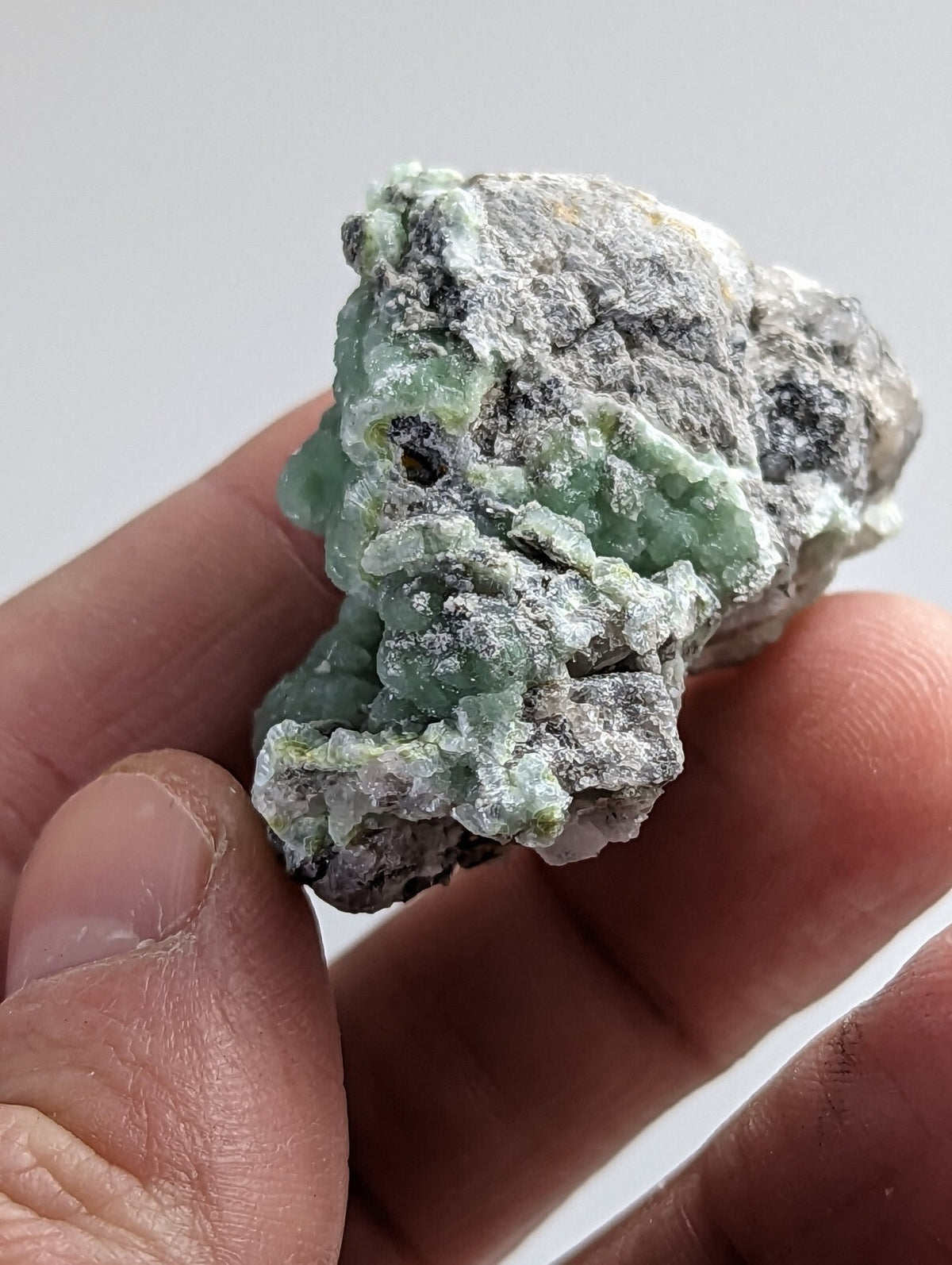 A rough, irregularly shaped mineral specimen with a mix of pale green and grayish-white hues, featuring a textured surface that appears to be composed of crystalline formations.