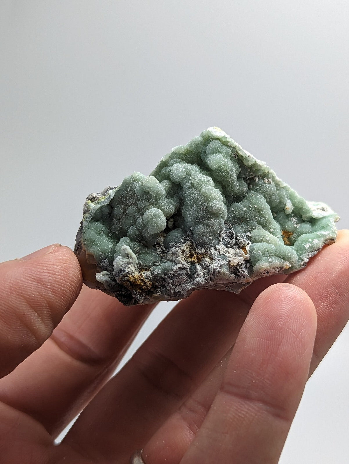 A hand holds a light green, textured mineral specimen with rounded, bubbly formations and patches of grayish matrix.