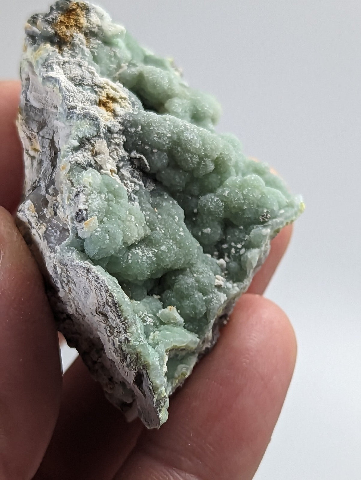 A hand holds a rough, light green mineral specimen with clustered crystalline formations and patches of white matrix.