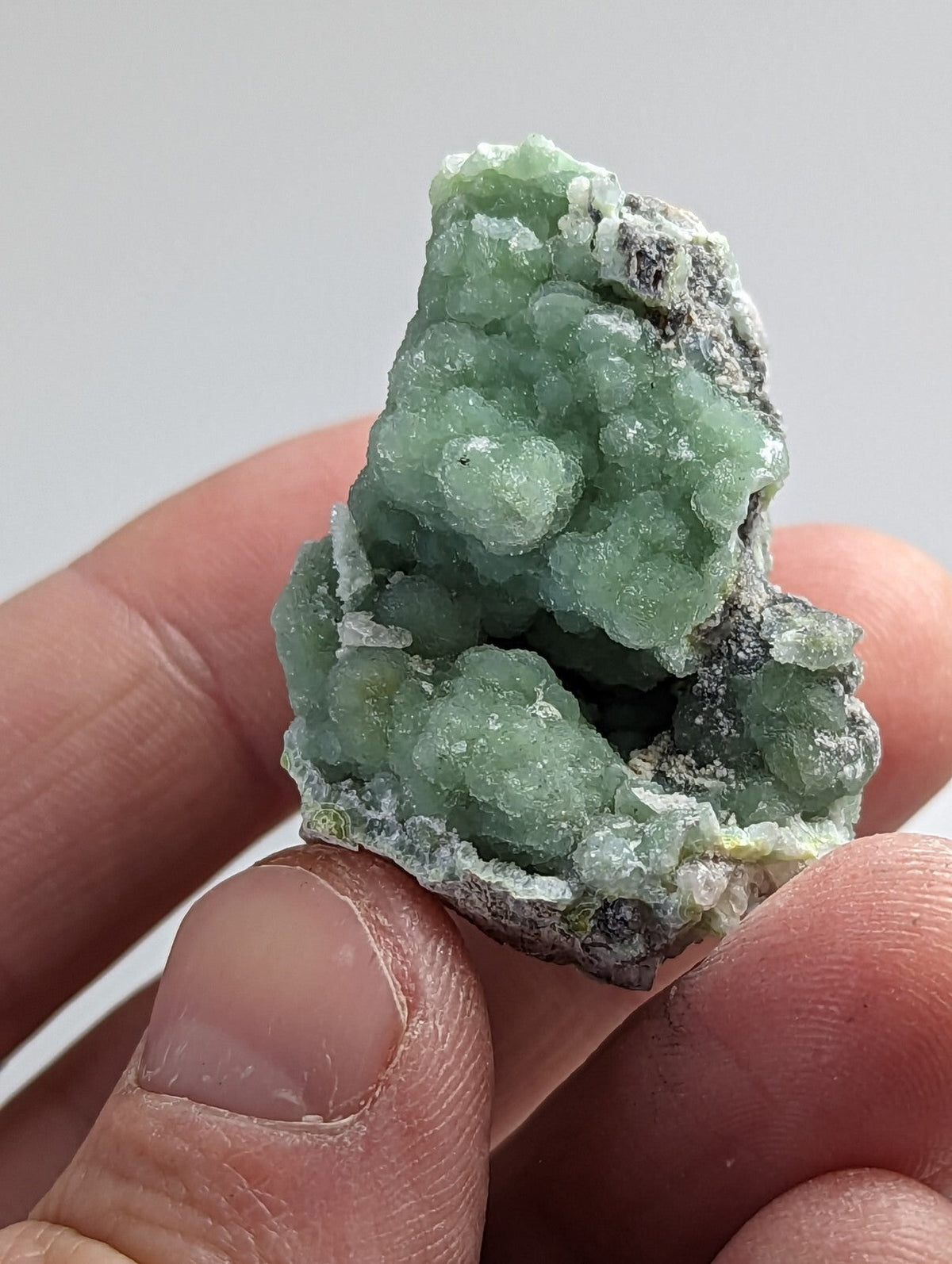 A hand holds a small, irregularly shaped mineral specimen featuring translucent green crystals clustered together with darker grayish matrix material.