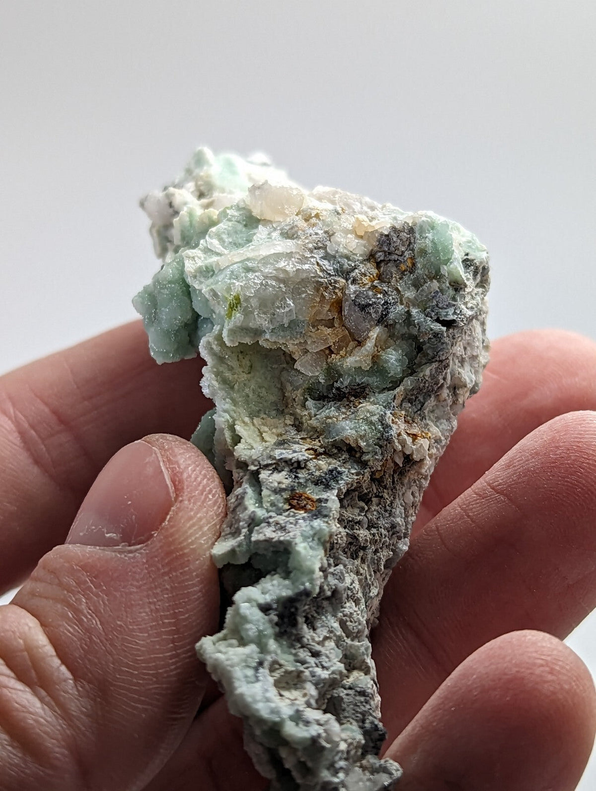 A hand holds a rough, irregularly shaped mineral specimen featuring patches of translucent light green crystals embedded in a darker gray matrix.