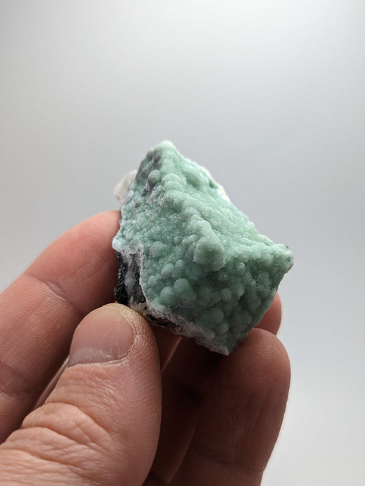 A hand holds a small, irregularly shaped piece of greenish-blue mineral with a textured surface featuring rounded nodules and patches of darker matrix.
