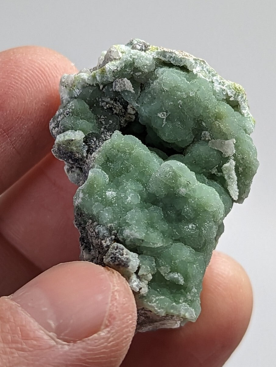 A hand holds a rough, green mineral specimen with crystalline formations and patches of grayish matrix.