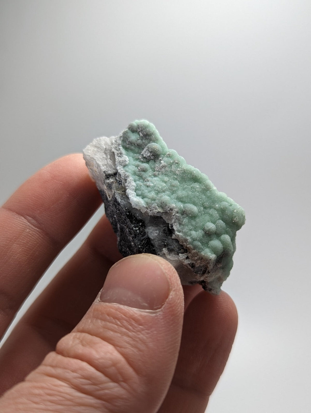 A hand holds a small, irregularly shaped specimen of greenish-blue chrysocolla with a textured surface and white crystalline patches.