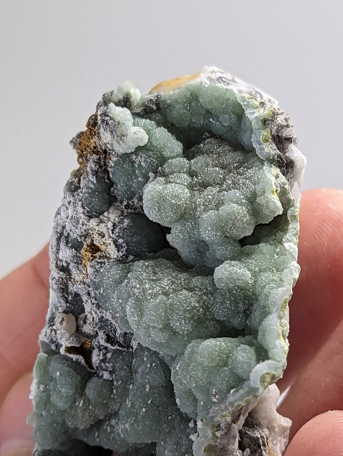 A hand holds a geode with vibrant green, bubbly crystal formations growing from dark gray rock.