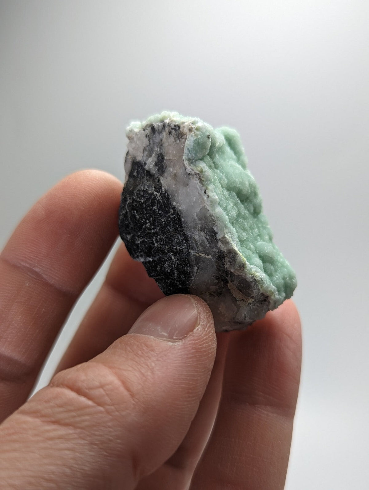 A hand holds a rough, irregularly shaped mineral specimen featuring vibrant green crystalline formations against dark black and white matrix material.
