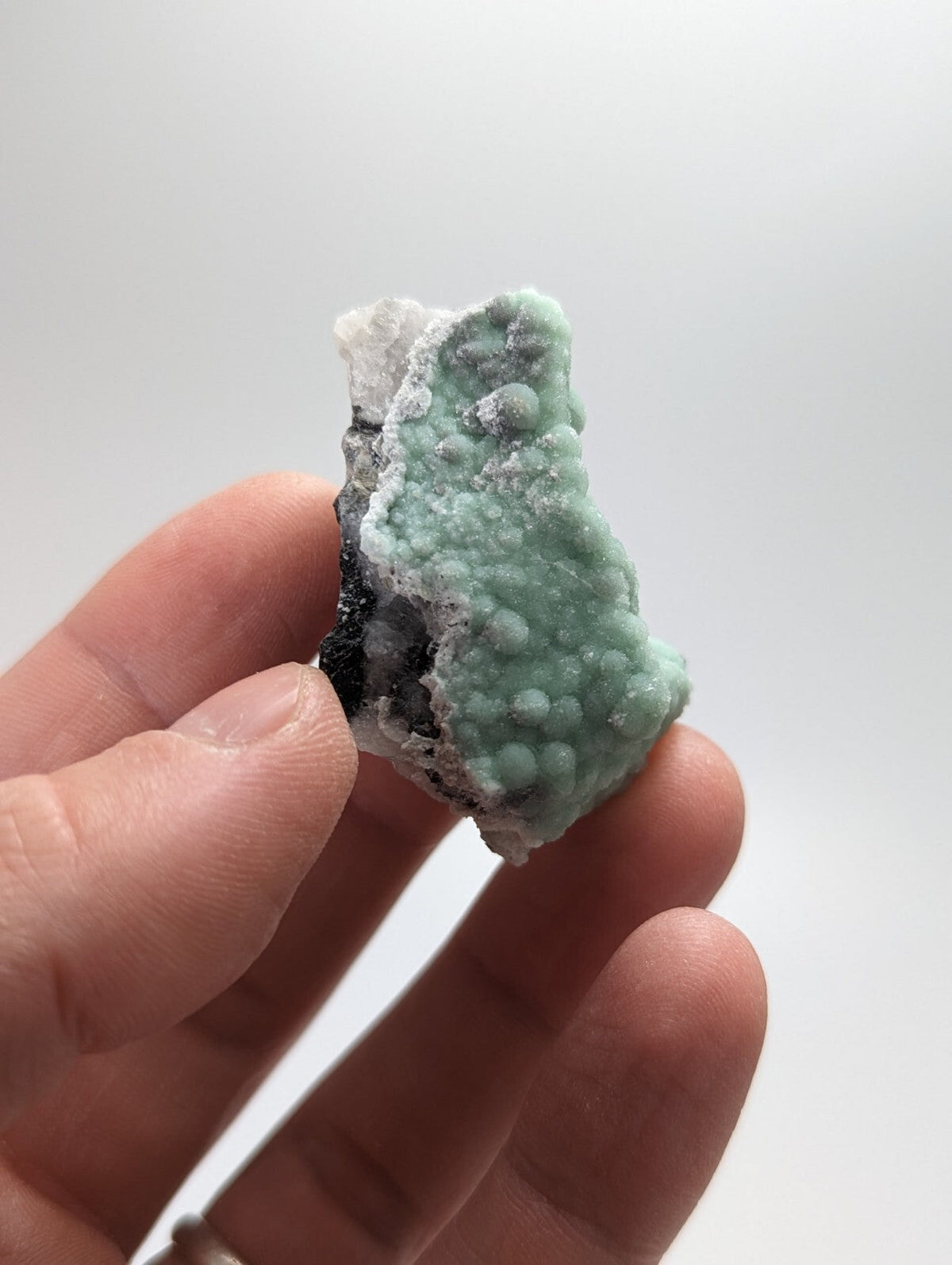 A hand holds a small, irregularly shaped piece of greenish-blue mineral with rounded nodules and white crystalline patches against a plain background.
