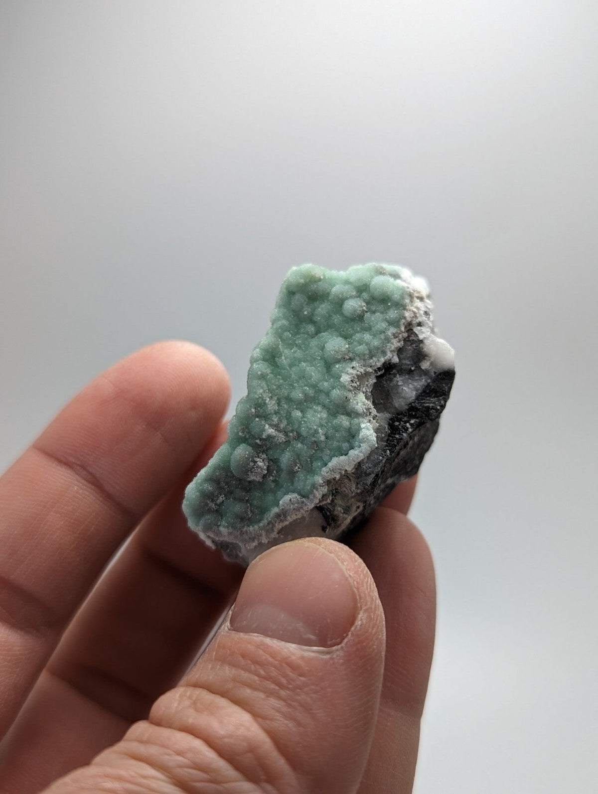 A hand holds a small, irregularly shaped mineral specimen featuring vibrant turquoise-green nodules with a rough, bumpy texture against a dark, contrasting base.