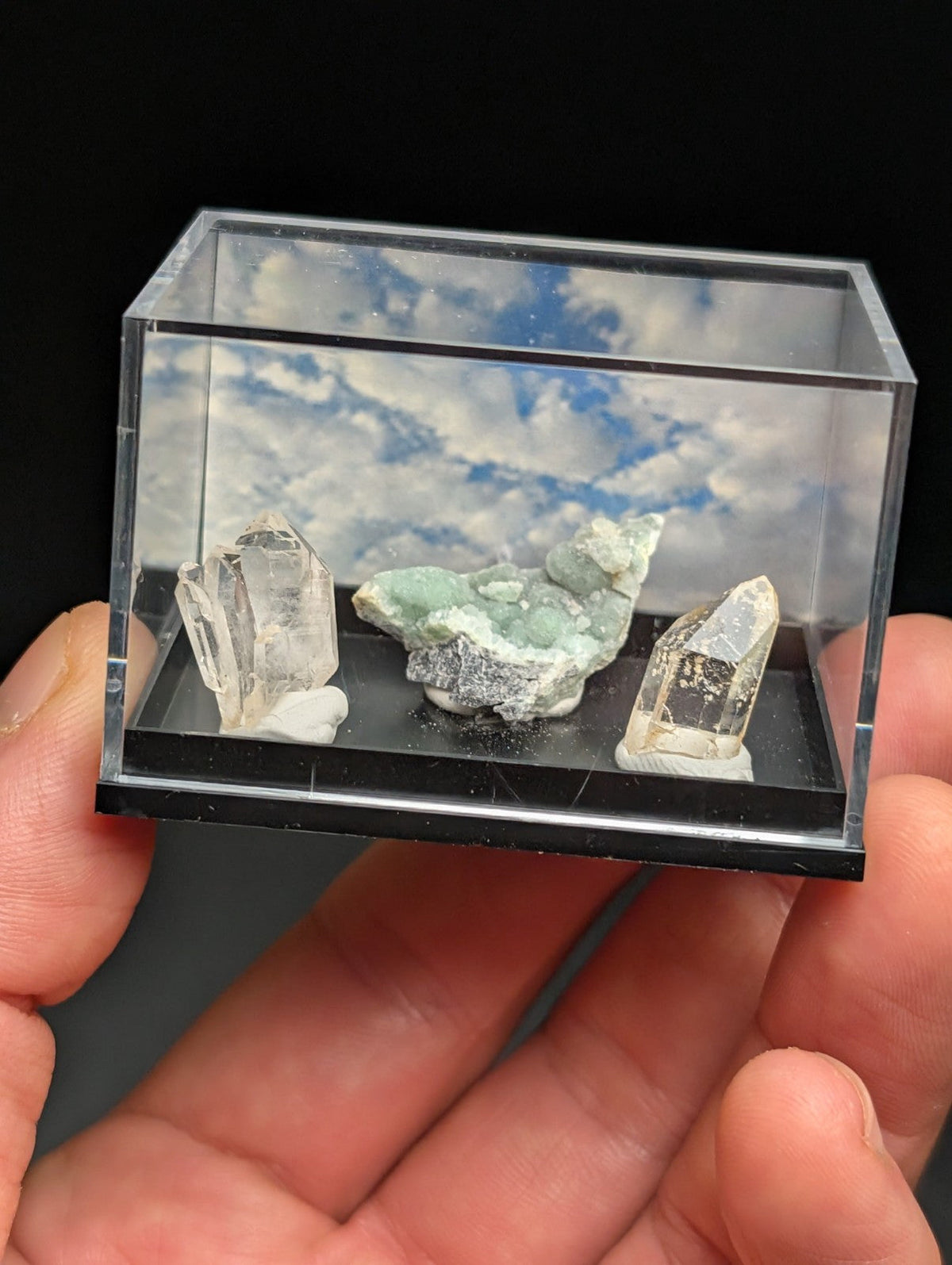 A clear acrylic display case holding three distinct mineral specimens: two transparent quartz crystals with pointed tips and a central greenish-blue mineral cluster, all set against a printed sky backdrop.