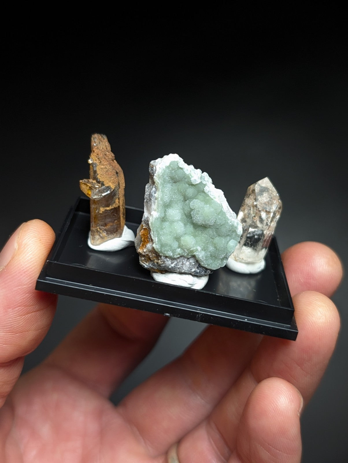 A hand holds a black display tray showcasing three distinct mineral specimens: one with brownish, translucent crystals; another featuring a vibrant green, clustered geode interior; and the third displaying clear quartz points.