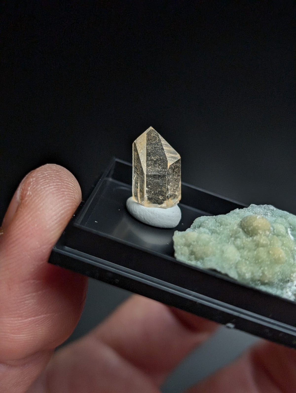 A translucent golden-yellow crystal with a faceted, geometric shape sits atop a small white pedestal within a black display tray.