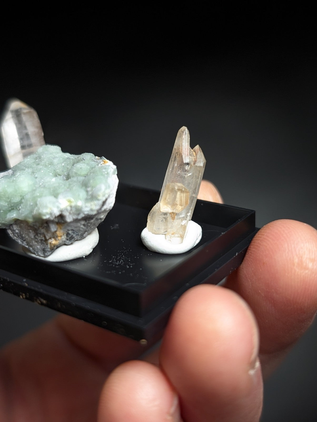 A clear quartz crystal with a pointed, faceted tip and subtle internal inclusions sits on a white circular base within a black display tray.
