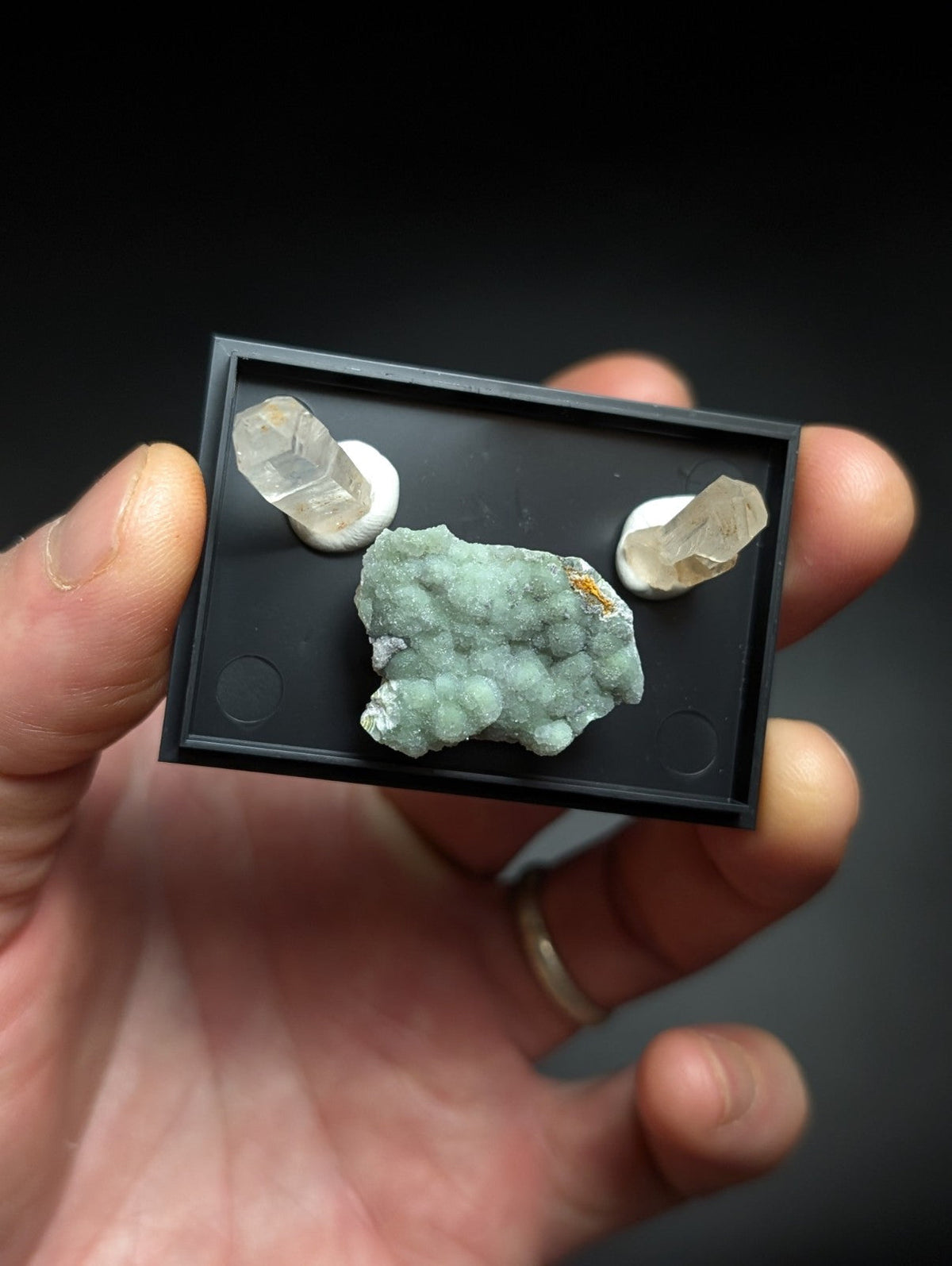 Black rectangular display case holding a greenish-blue mineral specimen and two clear quartz crystals.
