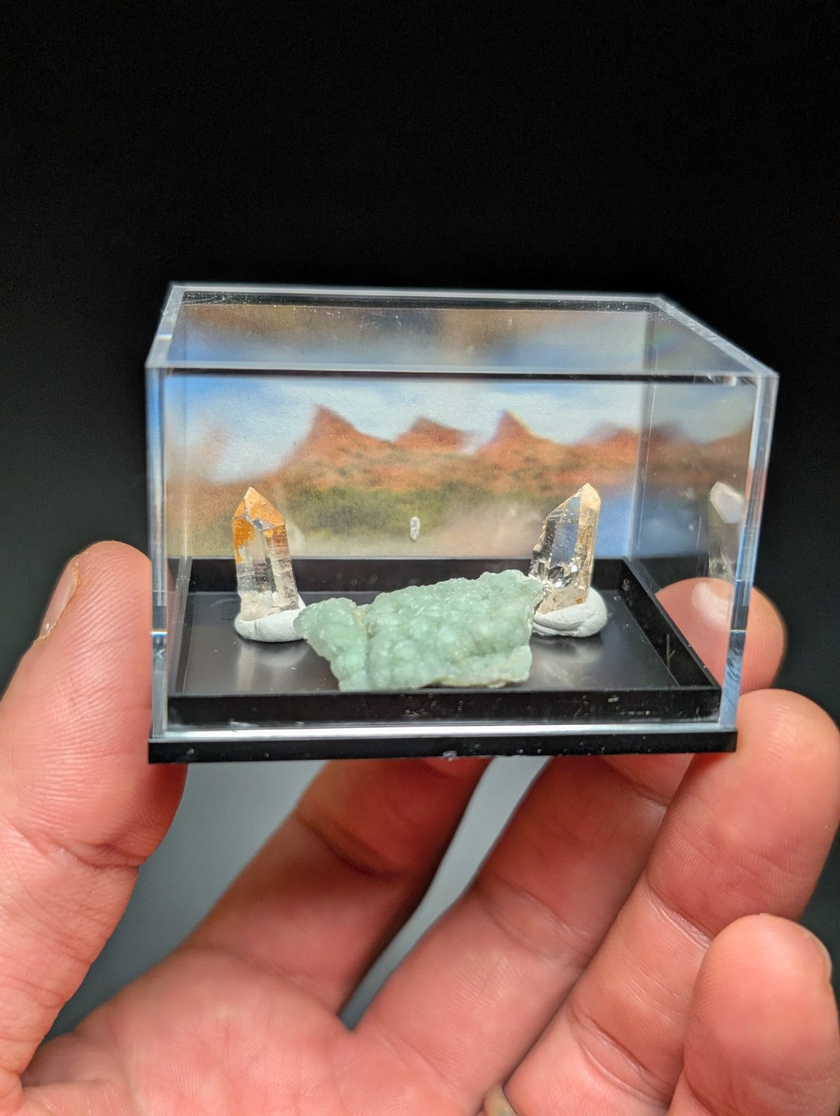 A clear acrylic display case holding a miniature desert landscape with two quartz crystals and a green mineral specimen.