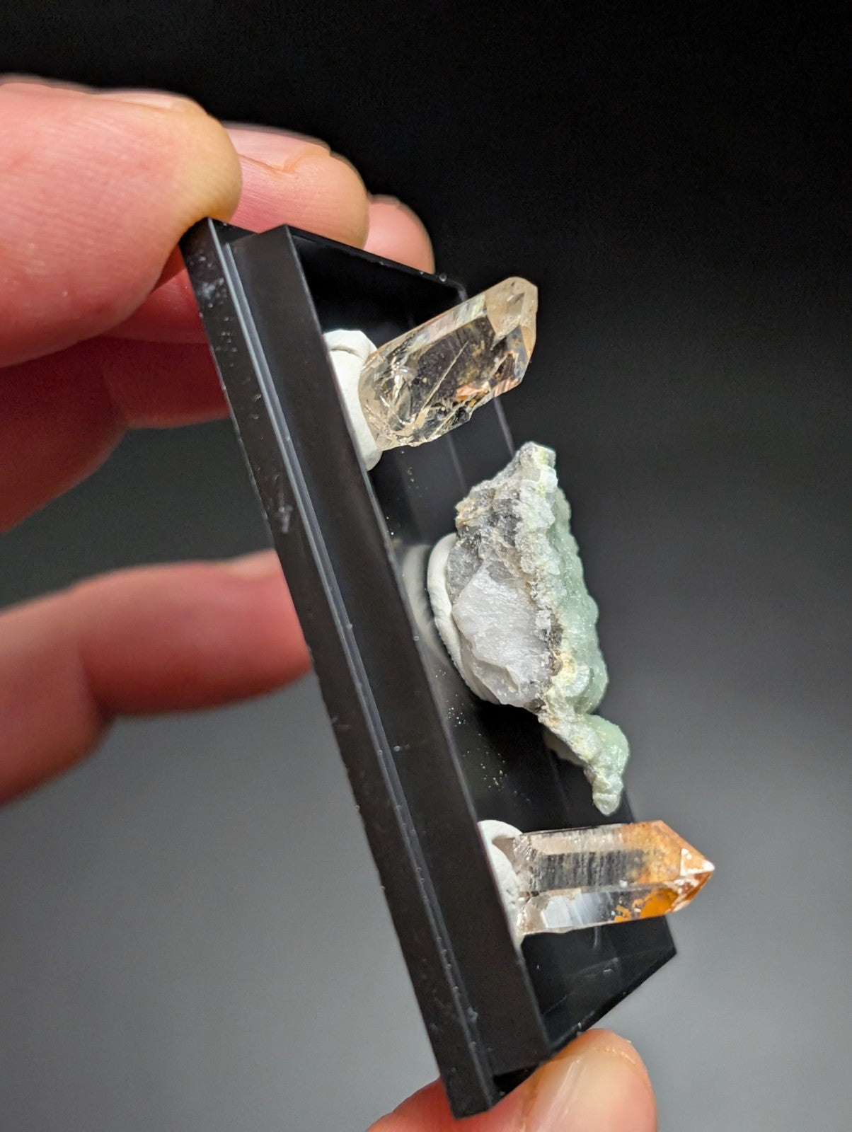 Black rectangular mineral display tray holding three distinct crystals: a clear quartz point, a greenish translucent specimen with white matrix, and an orange-tinted citrine crystal.