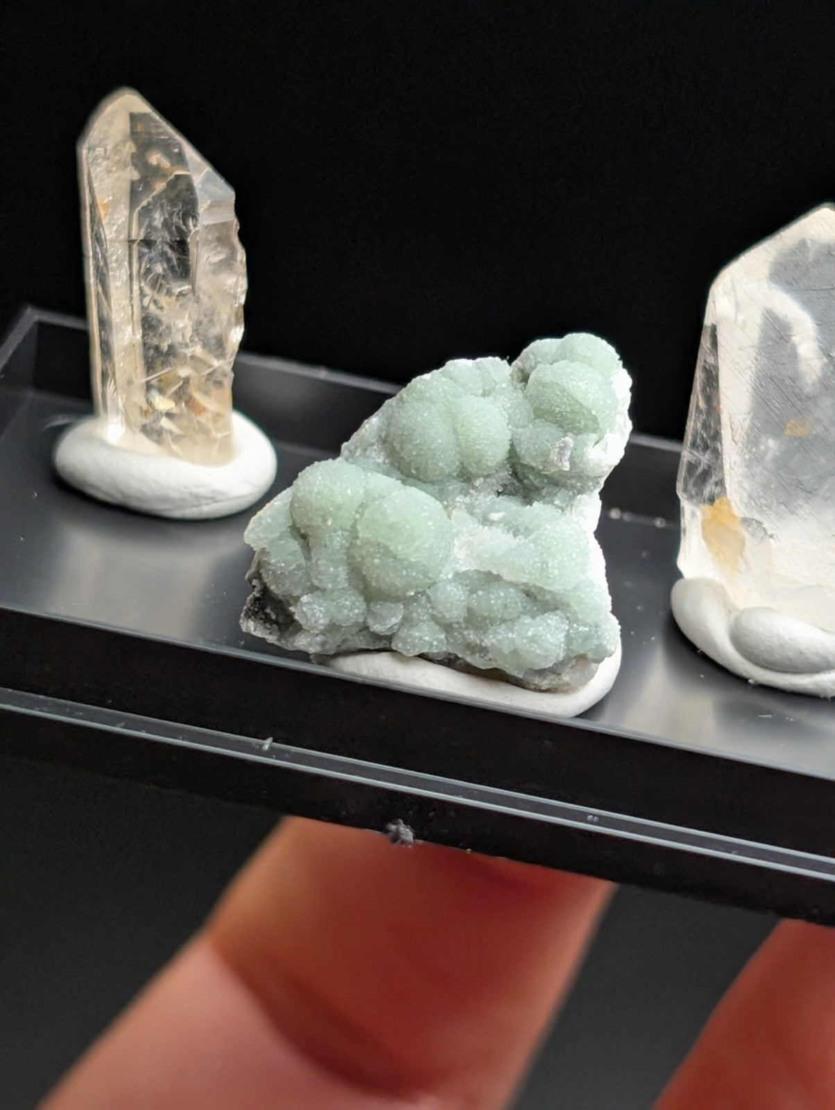 A cluster of light green, crystalline minerals with a textured, bumpy surface sits on a white base within a black display tray.