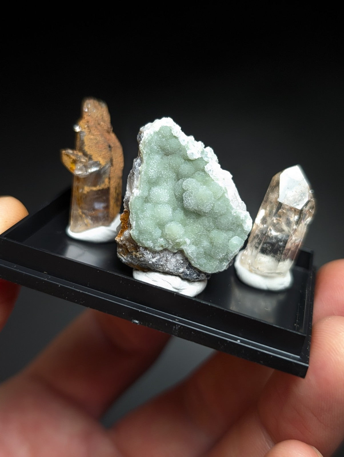 A black rectangular display tray holding three distinct mineral specimens: a translucent amber-colored crystal, a textured light green geode with white crystalline formations on its surface, and a clear quartz point.