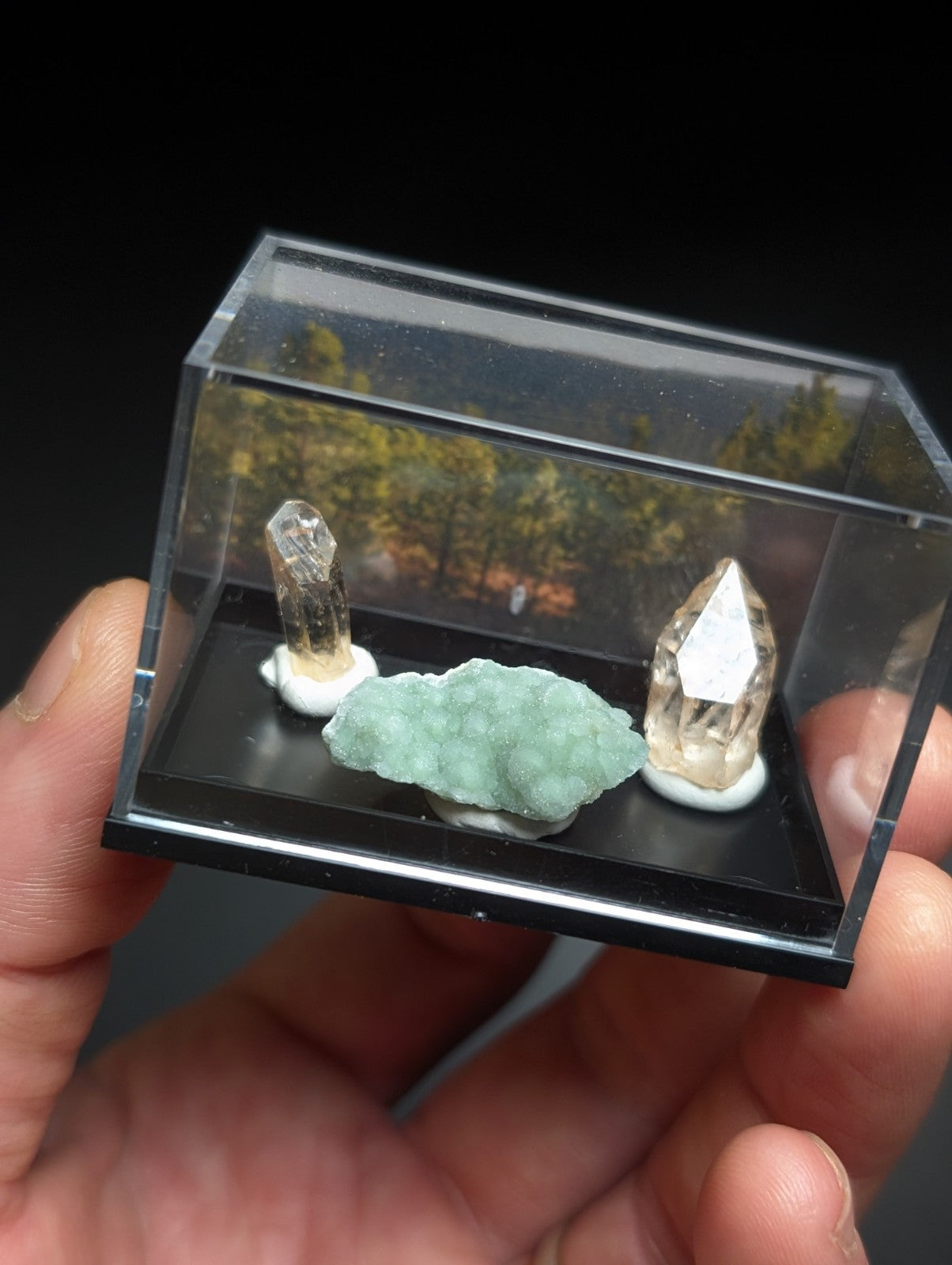 A clear acrylic display case holding three geodes and crystals, including a light green banded specimen with two translucent quartz points on white bases.
