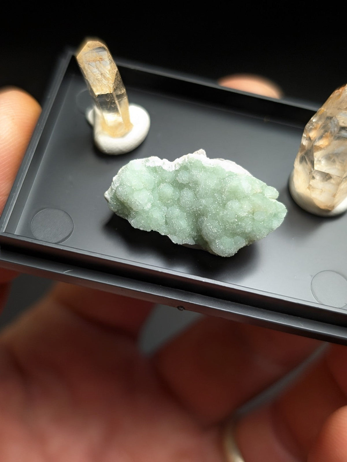 A pale green, crystalline mineral specimen with a textured, bumpy surface sits in the center of a black tray.