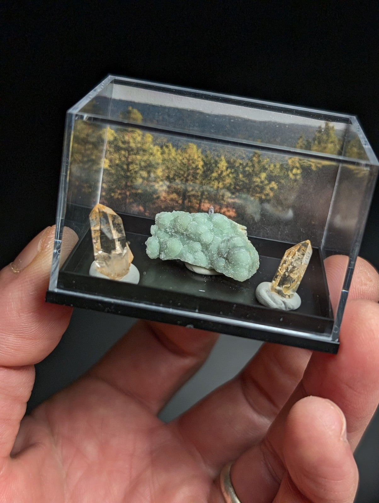 A clear acrylic display case holding three mineral specimens: a light green, textured geode-like rock and two translucent yellowish crystals with white bases.