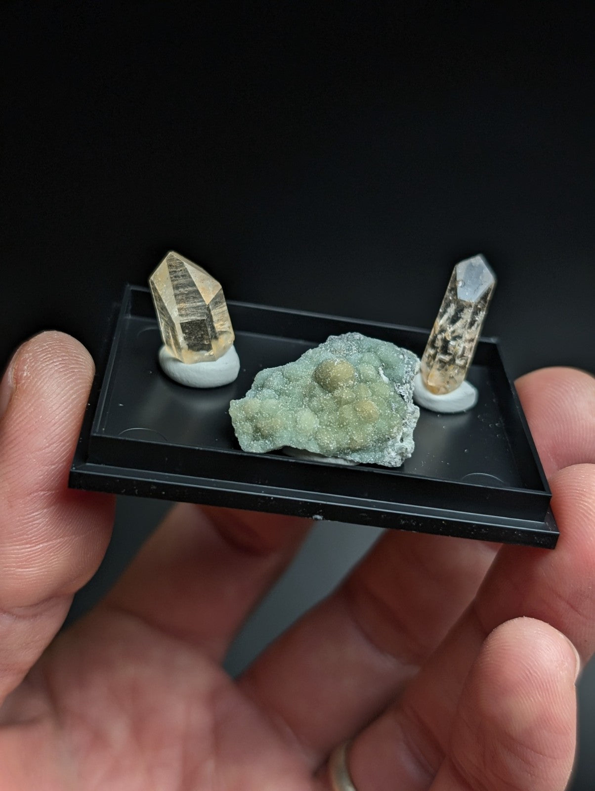 Black display tray holding two golden-yellow quartz crystals and a greenish mineral specimen.