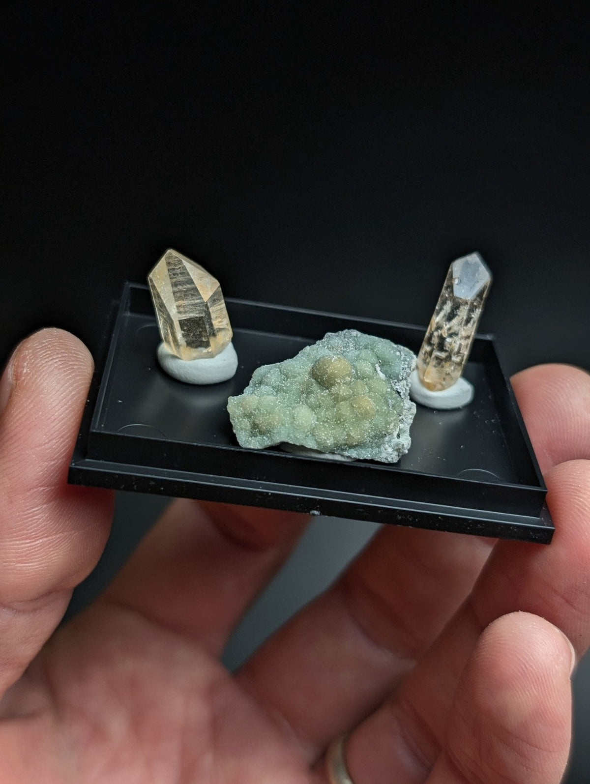 Black display tray holding two golden-yellow quartz crystals and a greenish mineral specimen.