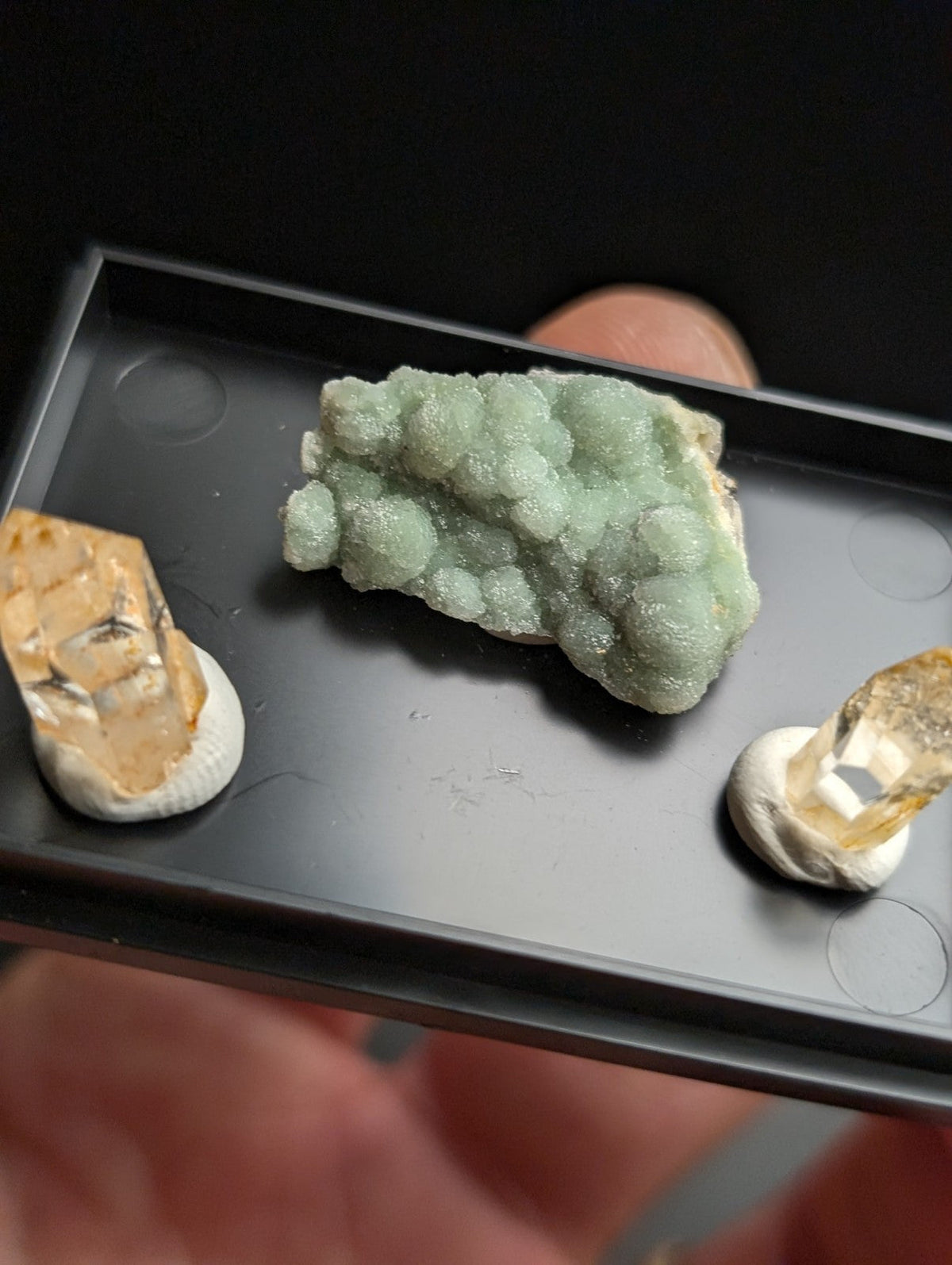 A light green, bubbly mineral specimen with a crystalline texture rests in the center of a black tray.