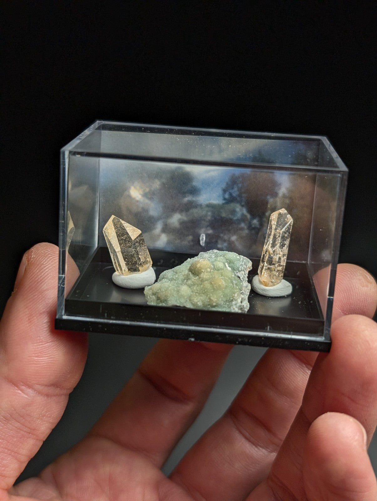 Clear acrylic display case holding two golden-brown quartz crystals and a greenish mineral specimen on white circular bases