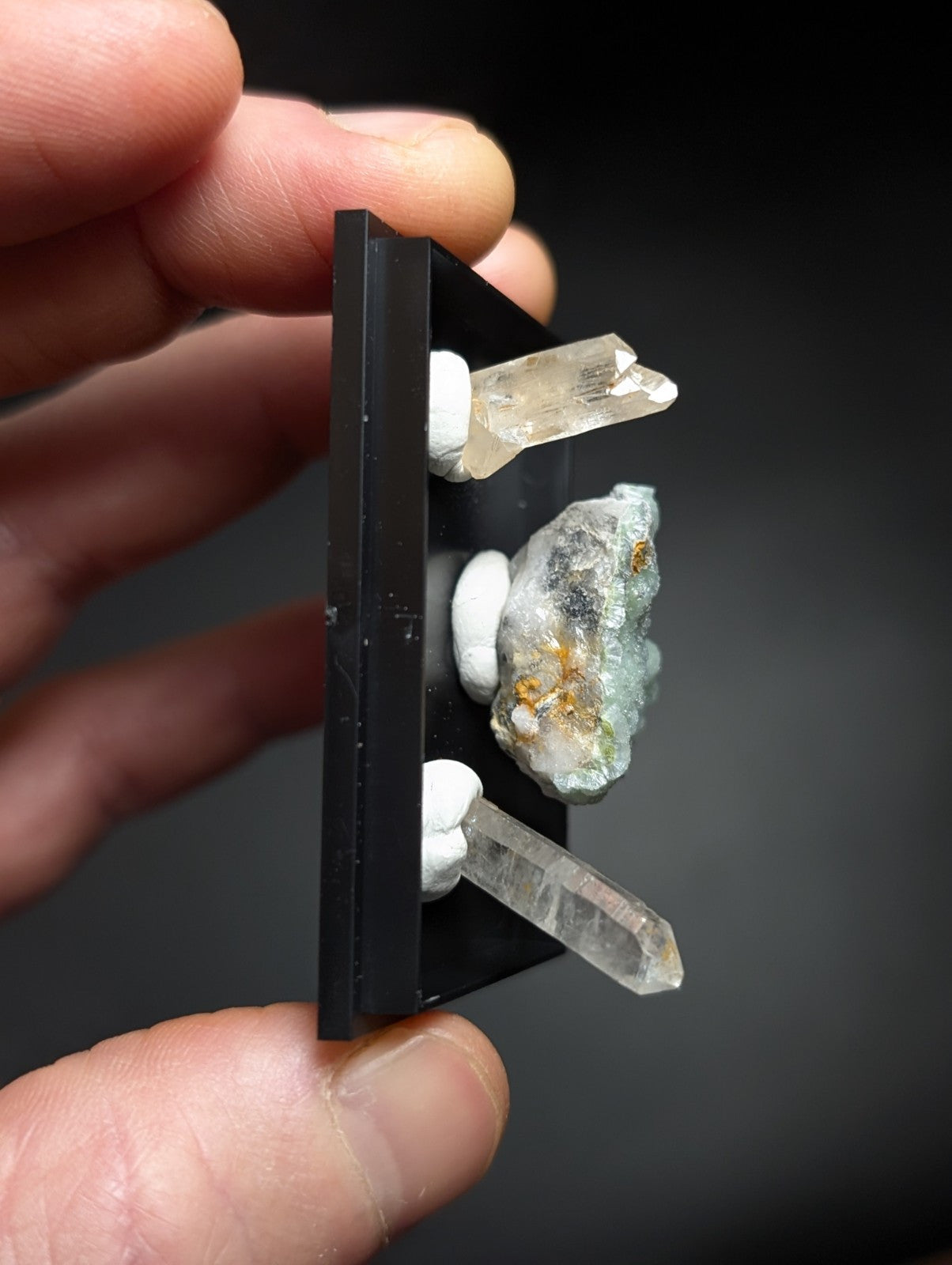 A black rectangular display stand holding three clear quartz crystals and one greenish mineral specimen with golden inclusions.