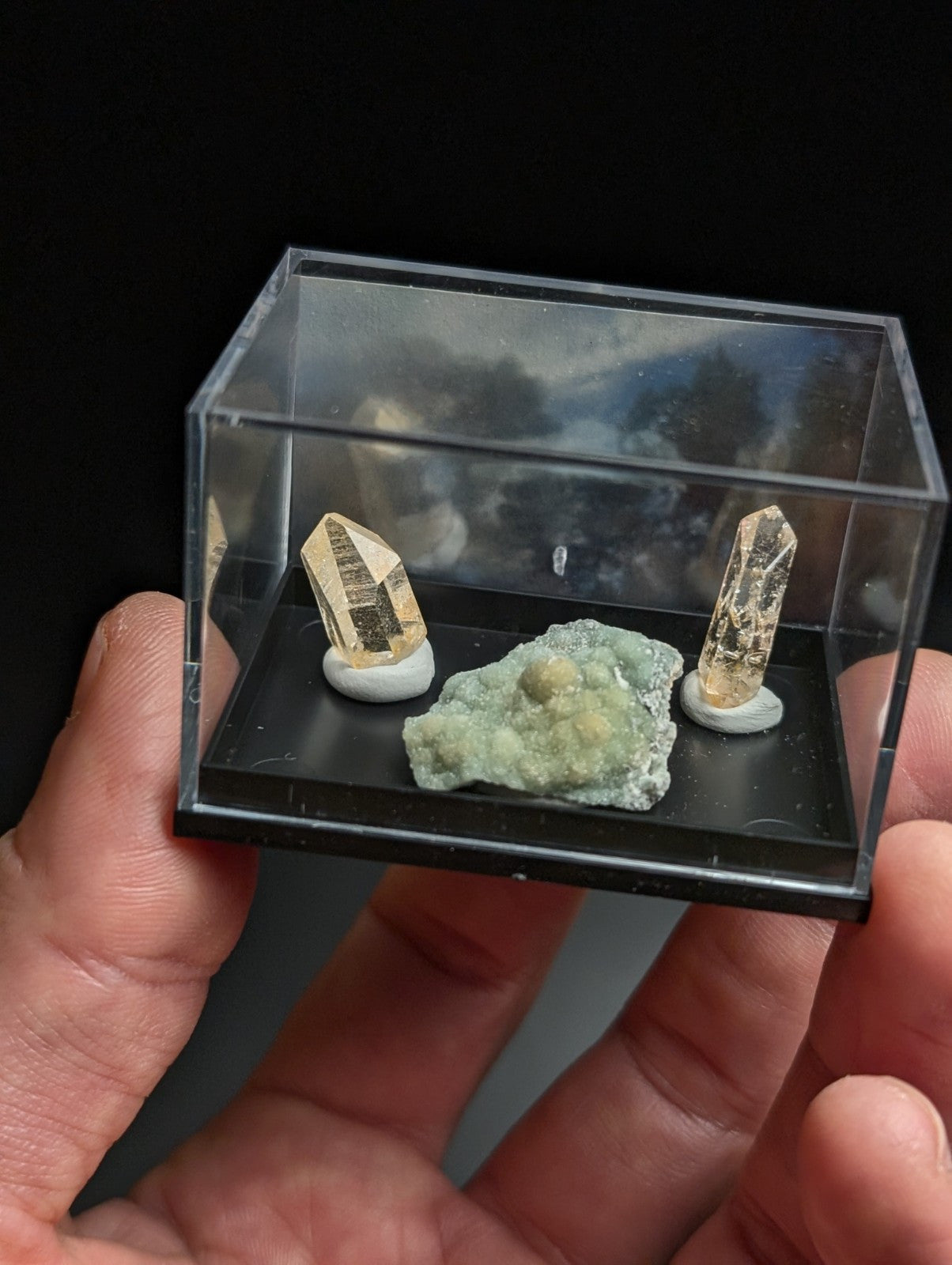 A clear acrylic display case holding two golden quartz crystals and a greenish mineral specimen on a black base.