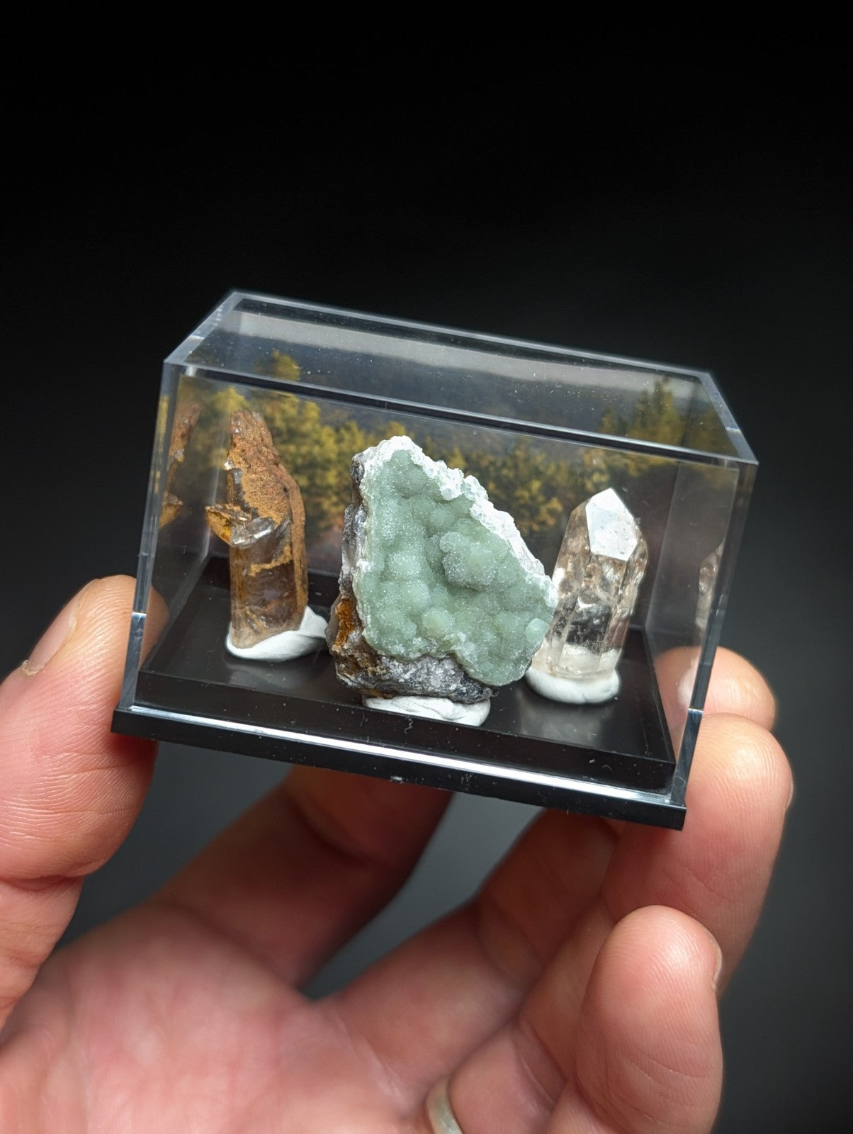 A clear acrylic display case holding three distinct mineral specimens: a brownish-orange crystal, a greenish-blue druzy formation with white crystalline coating on its base and sides, and a transparent quartz point.