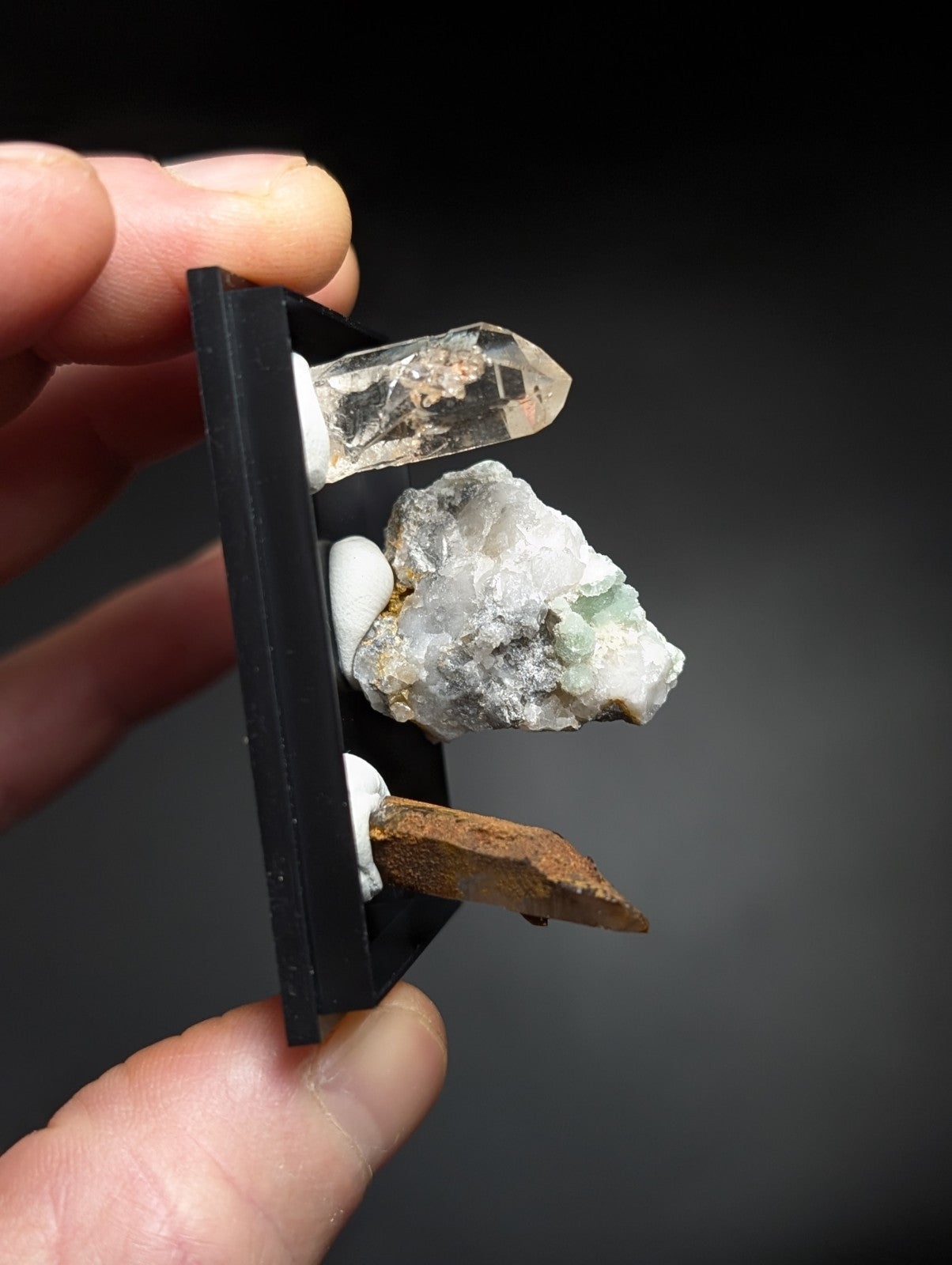 A black display stand holding a clear quartz crystal, a white and green mineral specimen with crystalline texture, and a brownish rock fragment.