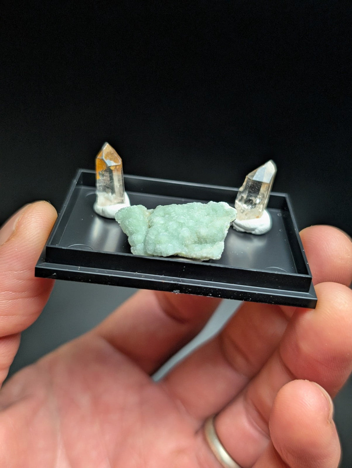 A black rectangular display tray holding a light green crystalline mineral specimen flanked by two clear quartz crystals on white bases.
