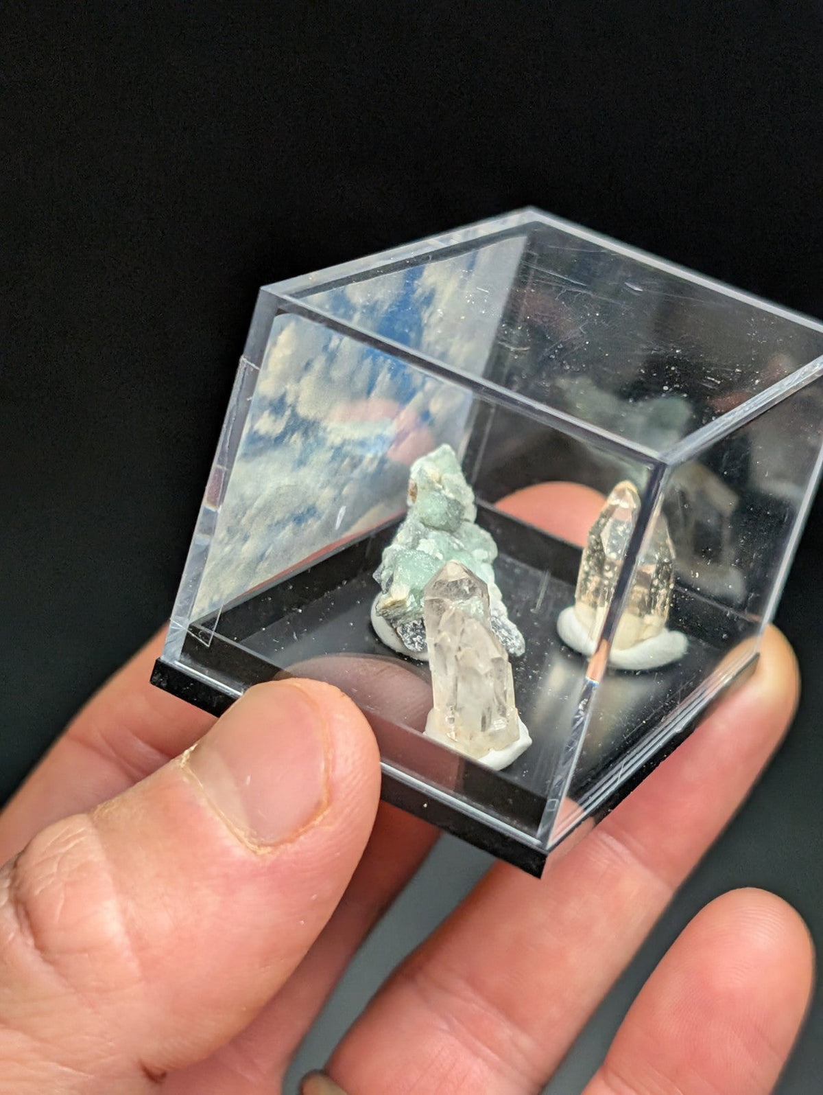 A clear acrylic display case holding two small, naturally formed mineral specimens with translucent green and white crystalline structures.